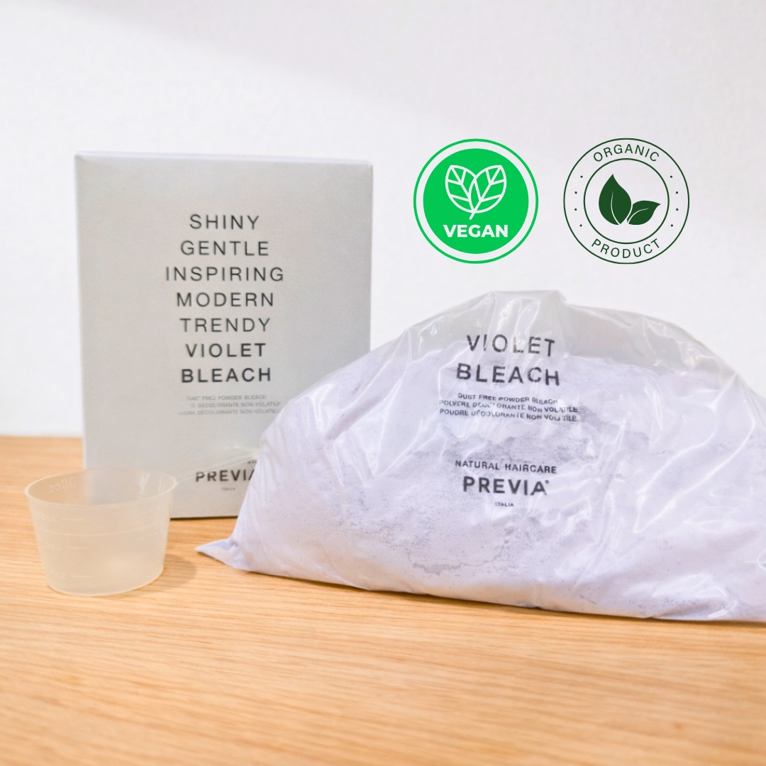 Embalagem de pó descolorante da Previa Italia sobre uma mesa de madeira. A imagem mostra um saco transparente com o Previa descoloração violeta, acompanhado pela sua caixa branca e um copo medidor. No canto superior direito, destacam-se os selos de produto vegan e orgânico. Ideal para obter um cabelo descolorado com tons frios, o produto é descrito como "Shiny, Gentle and Modern".